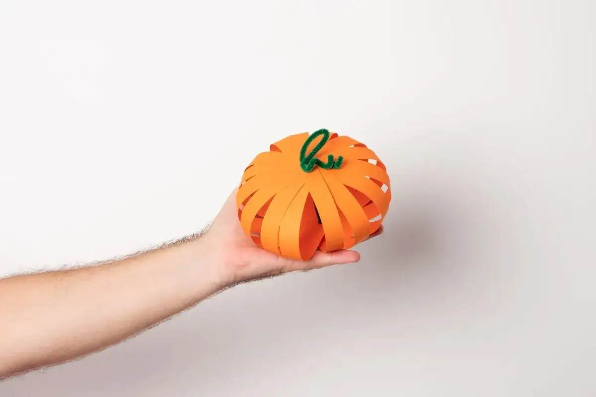 Ręcznie robione ozdoby na Halloween z papieru: pomarańczowa dynia z zieloną łodyżką, wykonana z pask&oacute;w papieru, trzymana na dłoni.