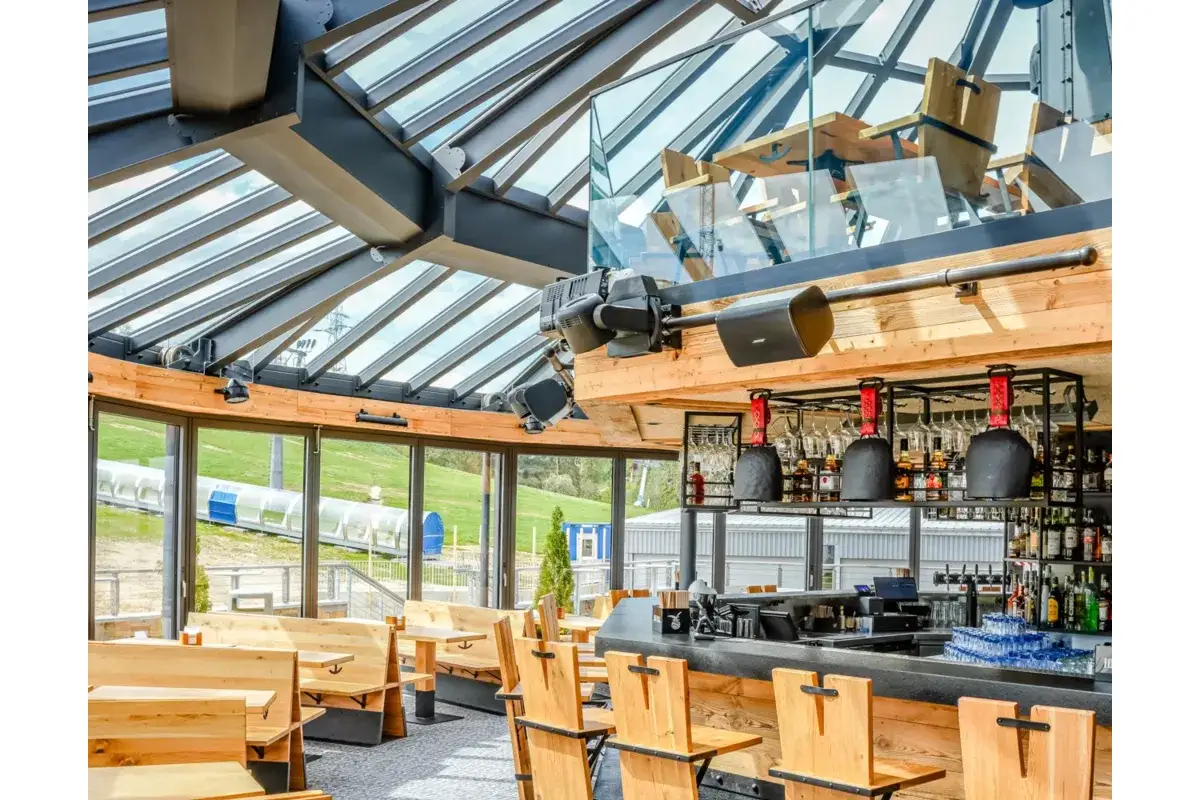 Nowoczesna restauracja w Bukowinie Tatrzańskiej z przeszklonym dachem i widokiem na stok. Drewniane ławy i bar tworzą przytulną atmosferę.