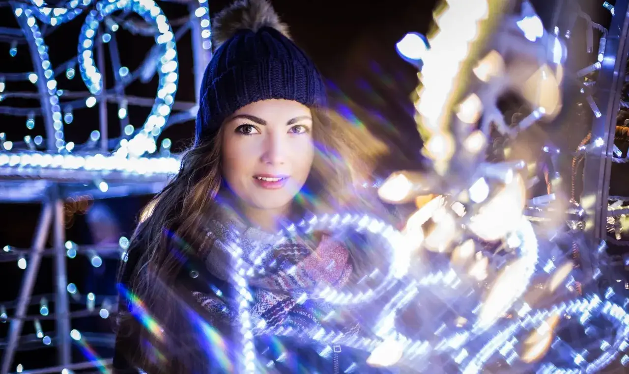 Kobieta w czapce i szaliku otoczona świątecznymi lampkami. Jasny obiektyw tworzy magiczne rozbłyski i bokeh.