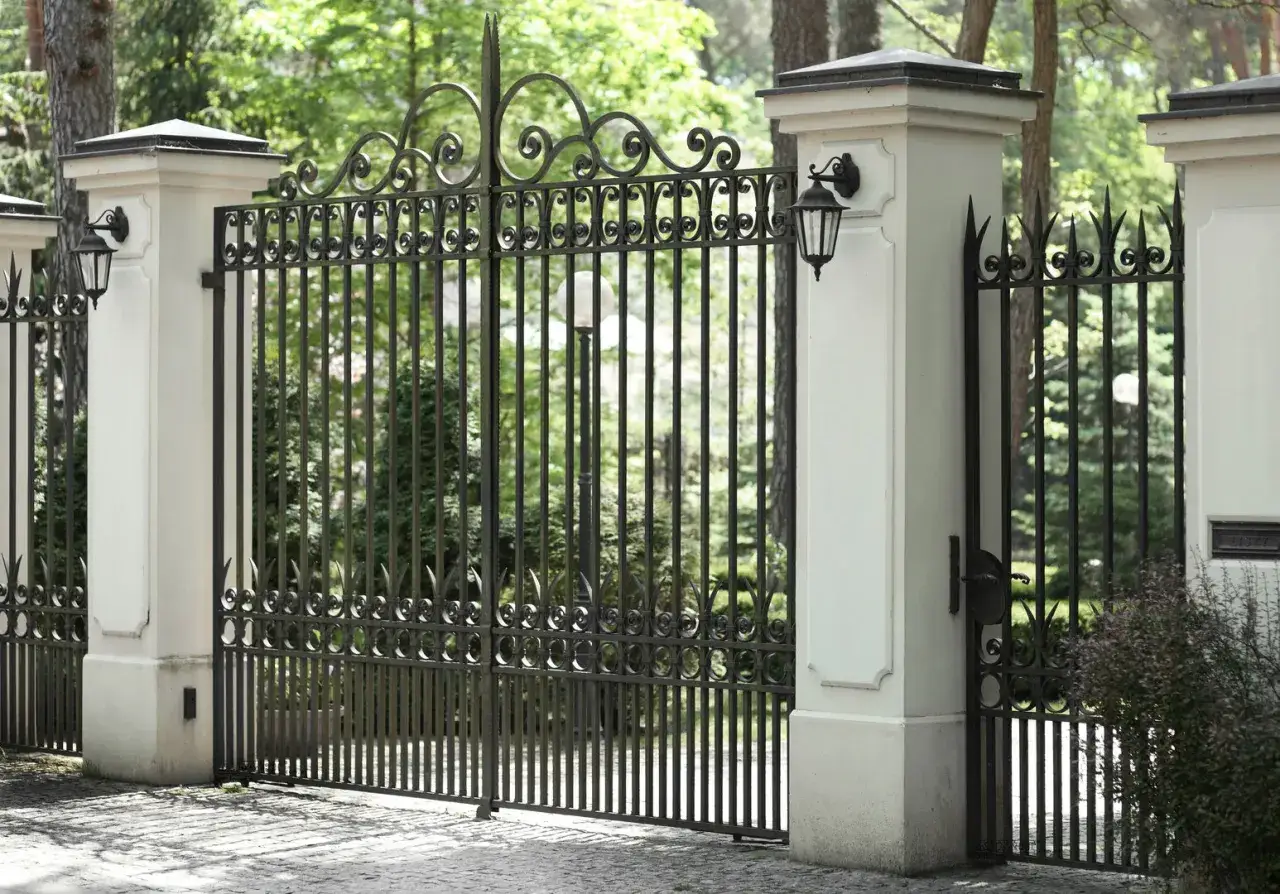 Elegancka, kuta brama z ozdobnymi wzorami, osadzona między białymi słupami z latarniami.