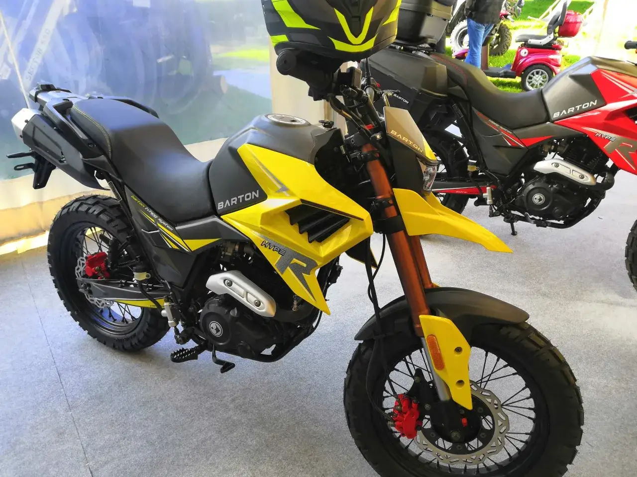 Ż&oacute;łty motocykl Barton Hyper 125, gotowy na zwiększenie mocy. Widoczny kask i inne motocykle w tle.