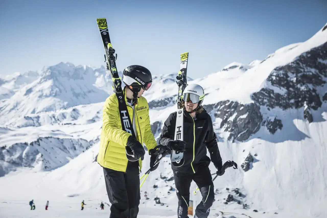 Dwóch narciarzy w kaskach i goglach idzie po ośnieżonym stoku, niosąc narty. Czy dłuższe narty są szybsze?
