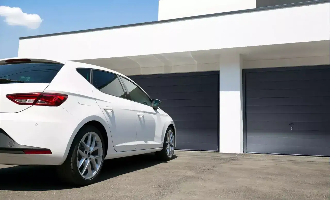 Nowoczesny dom z białym wykończeniem i dwoma ciemnoszarymi bramami garażowymi. Obok stoi biały samoch&oacute;d.