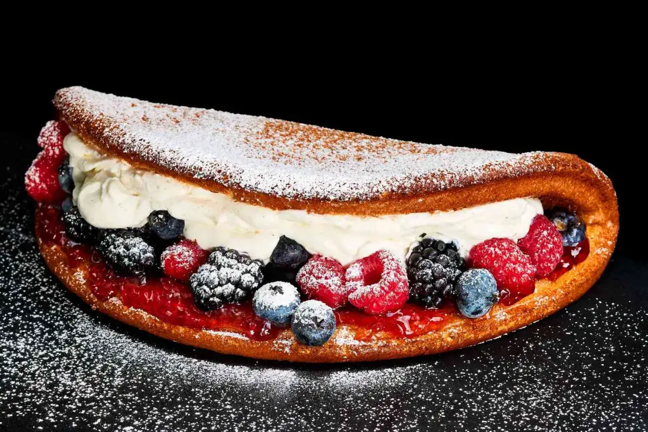 Puszysty omlet na słodko z bitą śmietaną, dżemem i świeżymi owocami leśnymi, posypany cukrem pudrem.