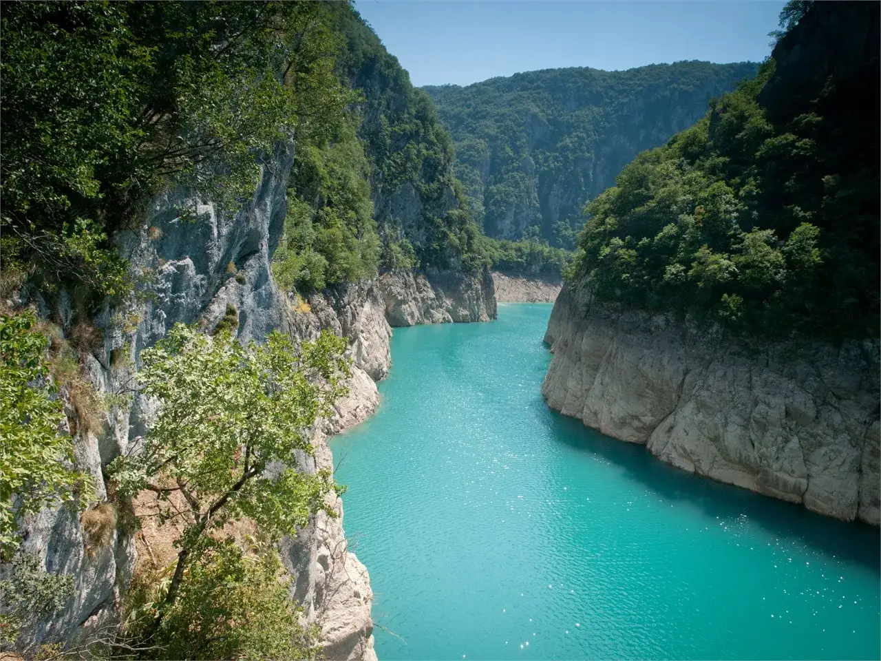 Turkusowe, krystalicznie czyste wody Pivsko Jezero otoczone stromymi, skalistymi zboczami porośniętymi bujną zielenią.