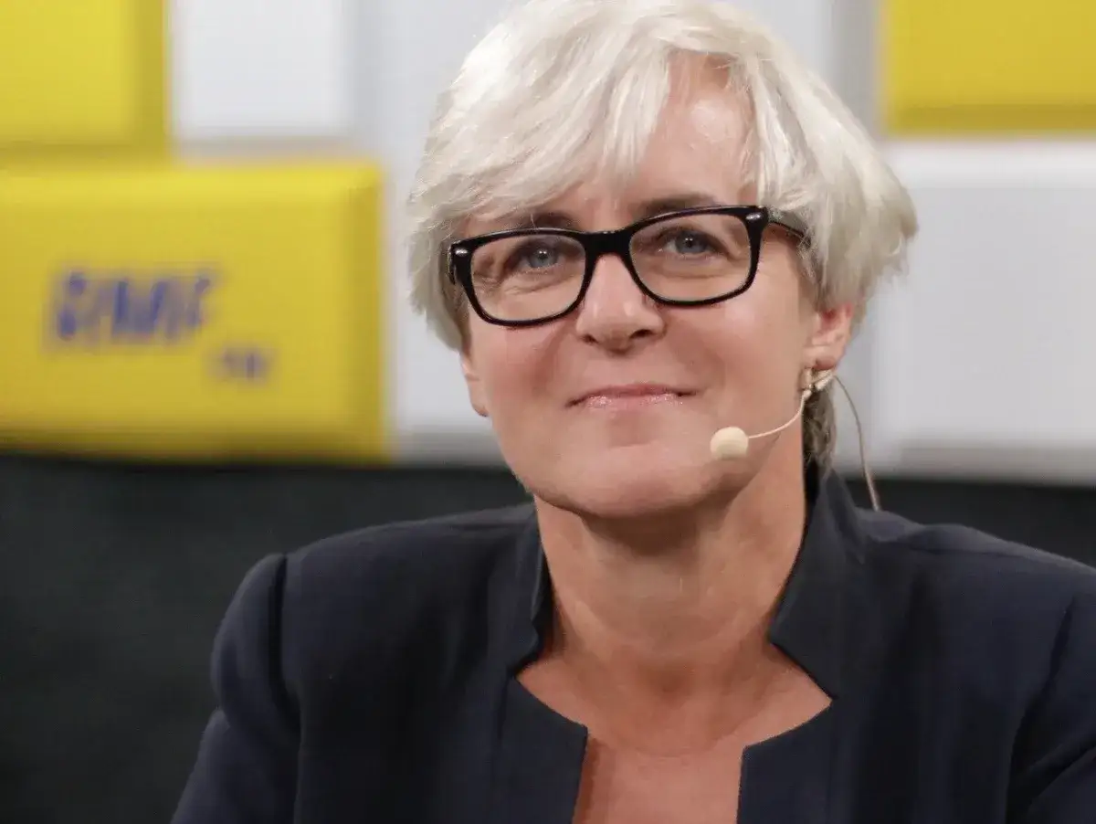 Joanna Kluzik Rostkowska w okularach, z mikrofonem przy uchu, uśmiecha się na tle żółtego baneru.