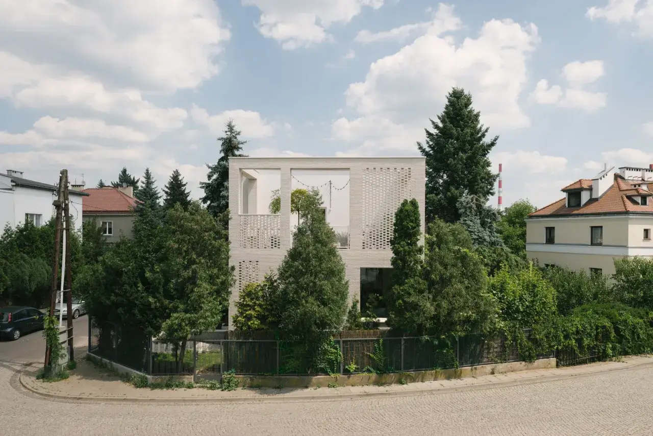 Nowoczesny dom kostka otoczony zielenią, z ażurowymi ścianami i dużymi oknami. Widać sąsiednie budynki i fragment ulicy.