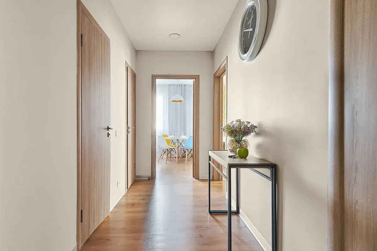 Jasny przedpok&oacute;j z drewnianymi drzwiami i podłogą. Na ścianie wisi zegar, a obok konsoli stoi wazon z kwiatami. Praktyczne ściany w przedpokoju dodają ciepła.