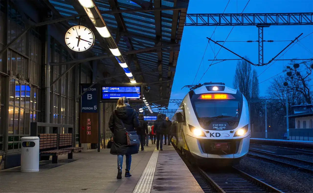 Na dworcu Wrocław Główny, tablica odjazdów pokazuje pociąg do Legnicy. Pasażerowie czekają na peronie 5.