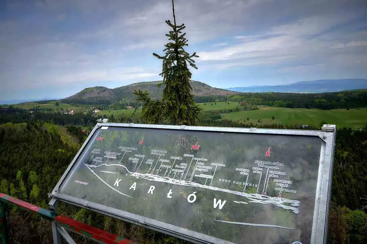 Widok z punktu widokowego na Karłów. Na pierwszym planie mapa z zaznaczonymi szczytami Gór Stołowych. W tle zielone wzgórza i lasy.