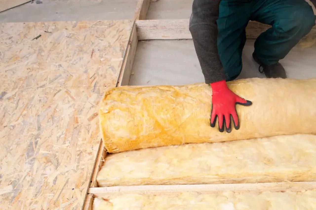 Rękawica w czerwonej rękawicy układa wełnę mineralną na płycie OSB na poddaszu.