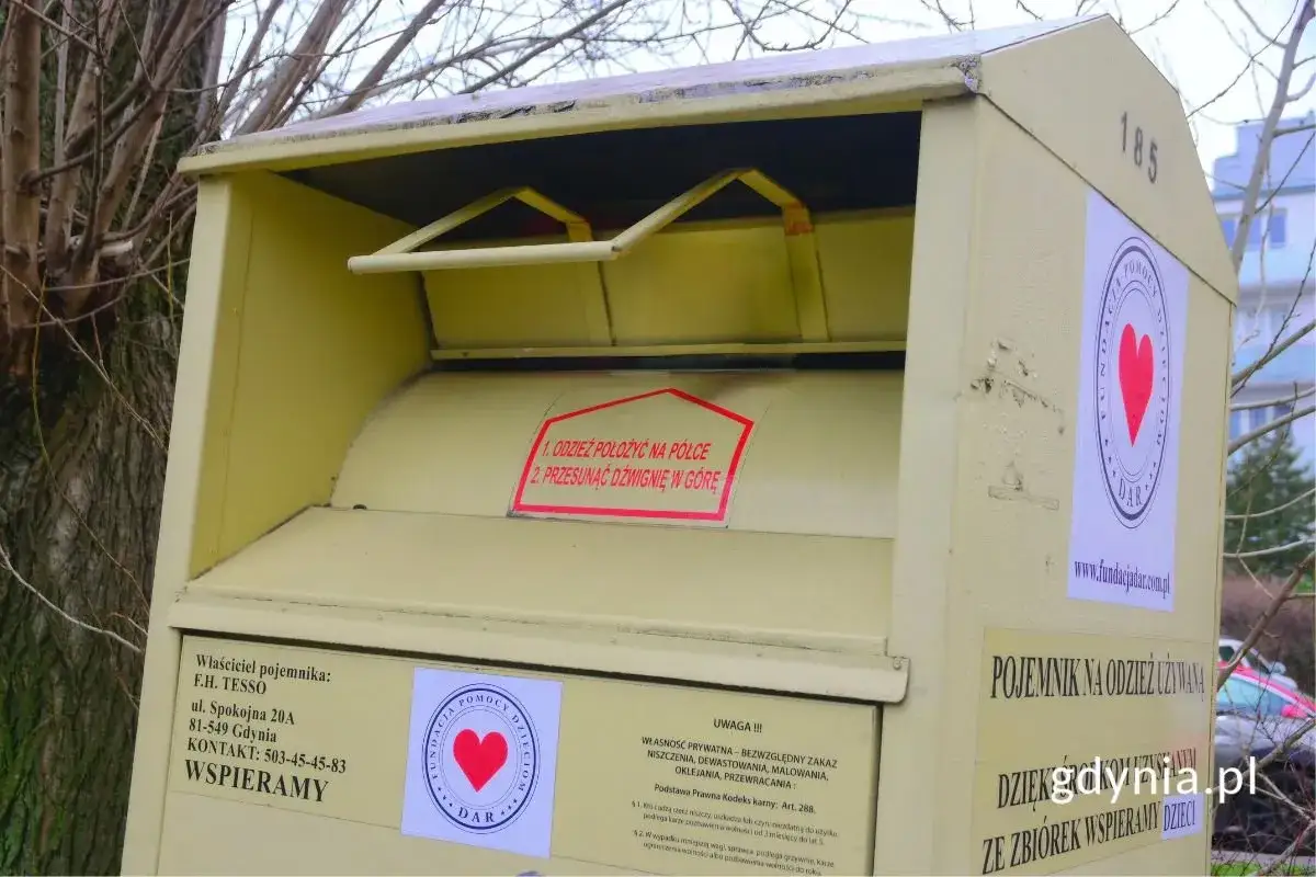 Żółte kontenery na ubrania stoją obok drzewa. Na jednym z nich widnieje napis 