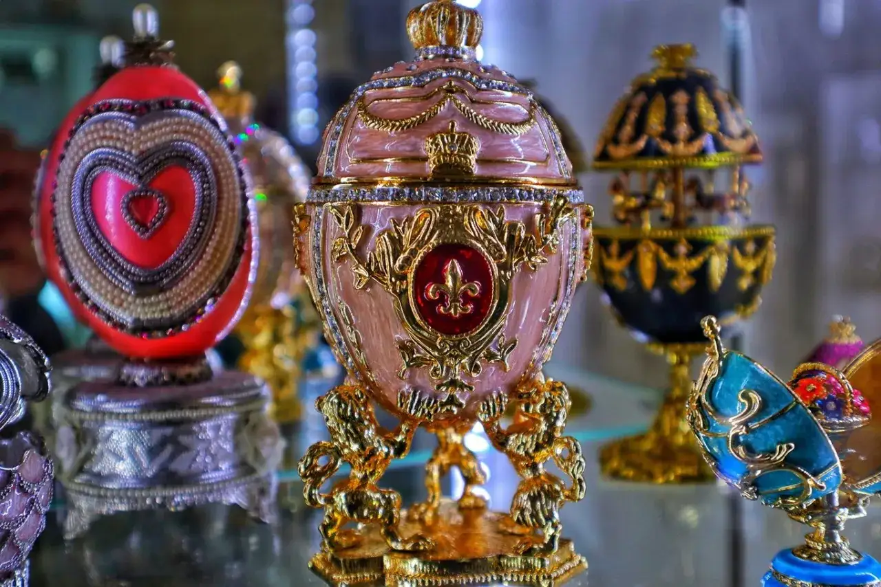 R&oacute;żowe jajko Faberge z koroną i złotymi lwami, obok czerwonego z sercem i czarnego z karuzelą.