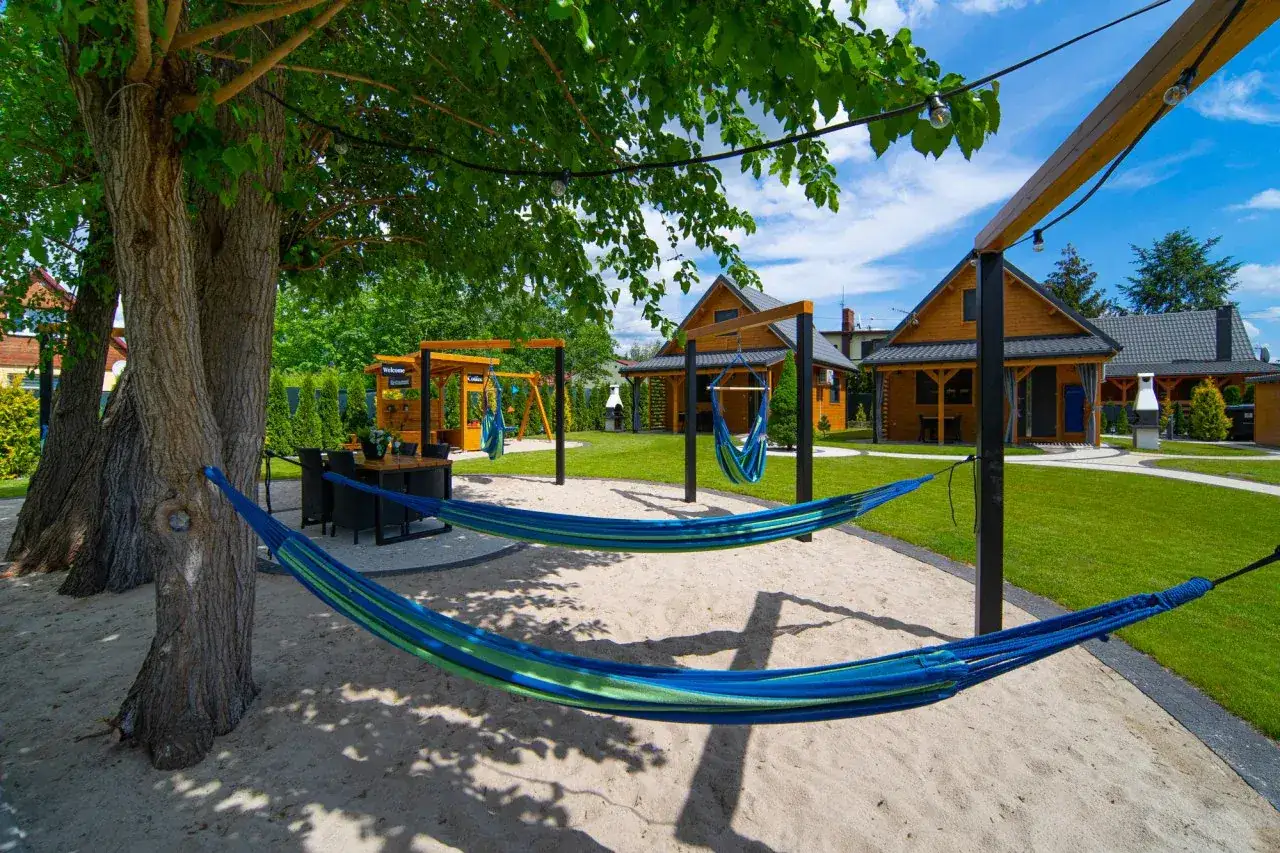 Relaks przy bacówkach nad jeziorem Goszcza. Dwie hamaki kuszą do odpoczynku na piaszczystym placu, w otoczeniu zieleni i drewnianych domków.