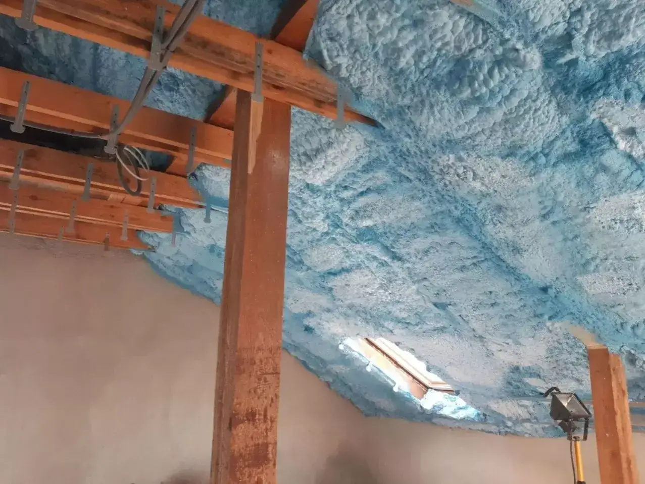 Izolacja pianką PUR na poddaszu to świetny sposób, jak ocieplić strych. Widać drewnianą konstrukcję dachu i niebieską piankę.