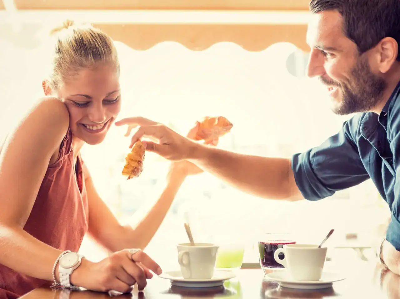 Uśmiechnięta kobieta przyjmuje kawałek rogalika od mężczyzny. Romantyczne triki działające na mężczyzn w kawiarni.