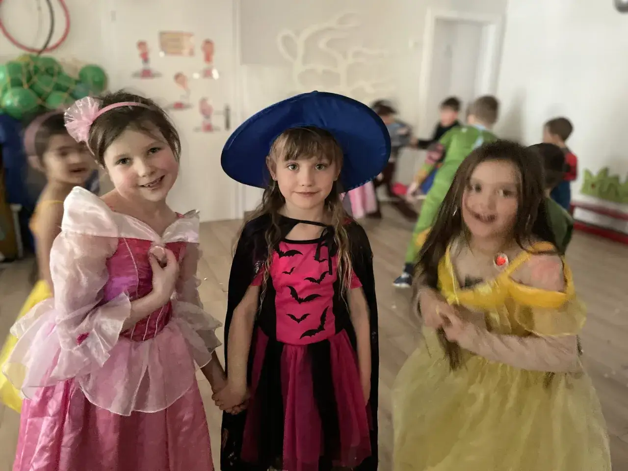 Trzy dziewczynki w przedszkolu świętują Andrzejki w strojach księżniczki, czarownicy i ż&oacute;łtej sukni. W tle widać inne dzieci.