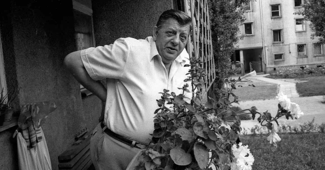 Mariusz Dmochowski, przed śmiercią, w białej koszuli, oparty o balustradę, z kwiatami w tle.