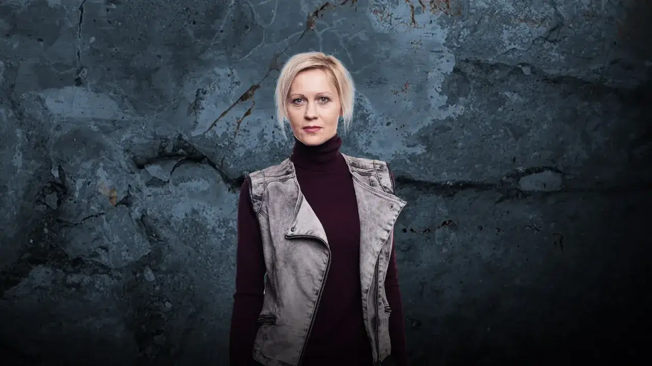 Lea Oleksiak, w bordowym golfie i szarej kamizelce, na tle surowej, pękającej ściany. Jej życie prywatne kryje się za tym spojrzeniem.