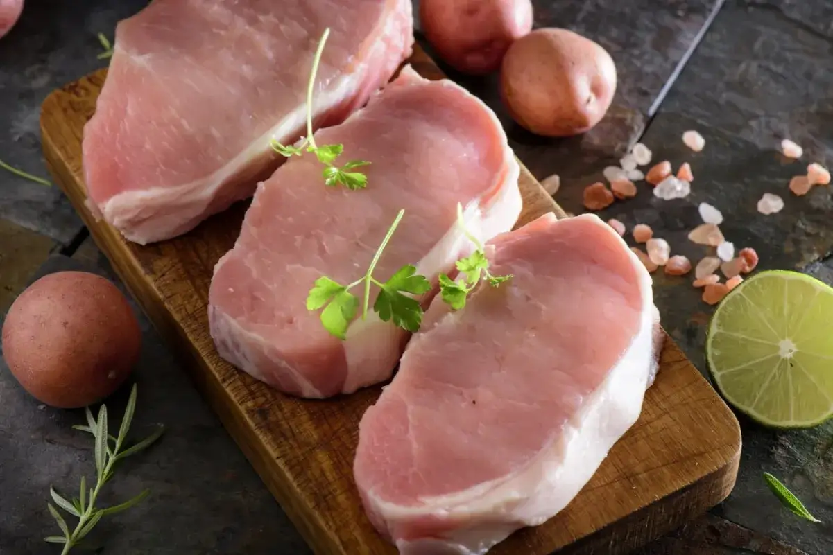 Surowe plastry schabu z grilla, udekorowane natką pietruszki, obok ziemniak&oacute;w, limonki i soli.