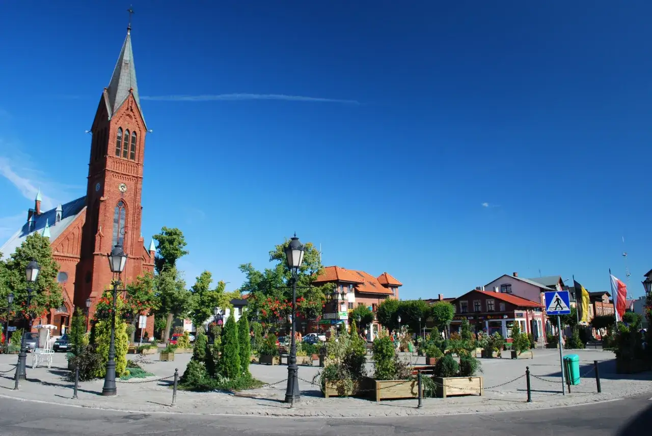 Rynek w Kartuzach z ceglanym kościołem i zielenią. Atrakcje turystyczne miasta zachęcają do spacer&oacute;w.