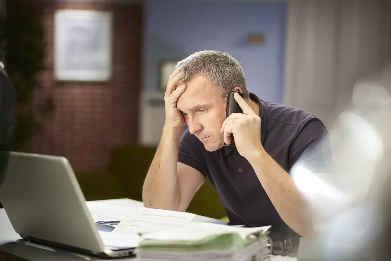 Zmartwiony mężczyzna rozmawia przez telefon, szukając informacji, jak odzyskać pieniądze z zamkniętego konta. Na biurku laptop i dokumenty.