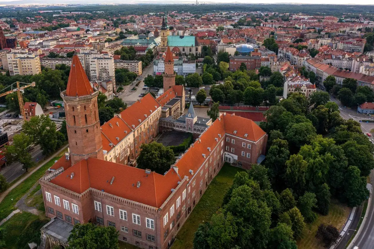 Widok z lotu ptaka na zamek w Legnicy, z charakterystyczną wieżą i czerwonymi dachami. W tle rozciąga się miasto, ukazując jego liczne atrakcje.