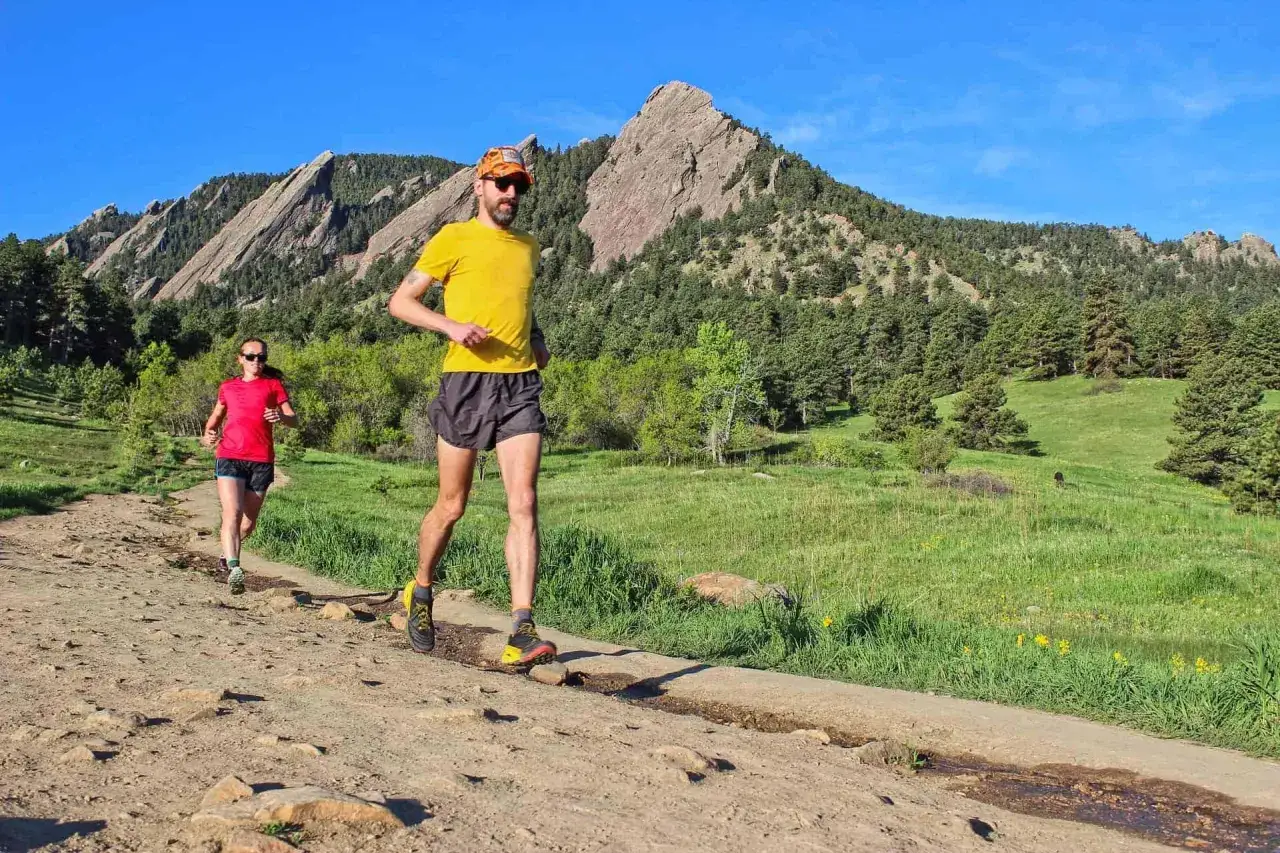 Dw&oacute;ch biegaczy pokonuje kamienisty szlak na tle g&oacute;rskich krajobraz&oacute;w. Słońce i piękna pogoda sprzyjają biegowi anglosaskiemu.