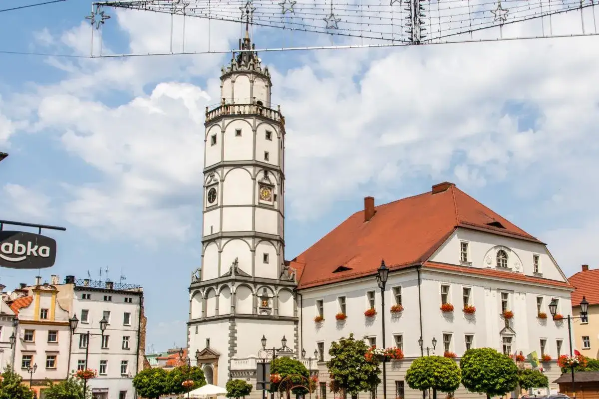 Wieża ratuszowa i biały budynek z czerwonym dachem to gł&oacute;wne paczk&oacute;w atrakcje. W tle błękitne niebo z chmurami.