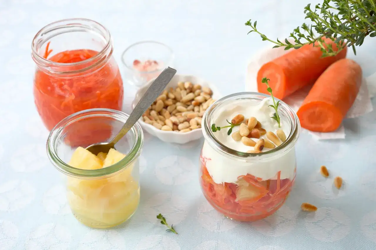 Słodki deser z marchewką i ananasem w słoiczku, ozdobiony jogurtem i orzeszkami pinii.