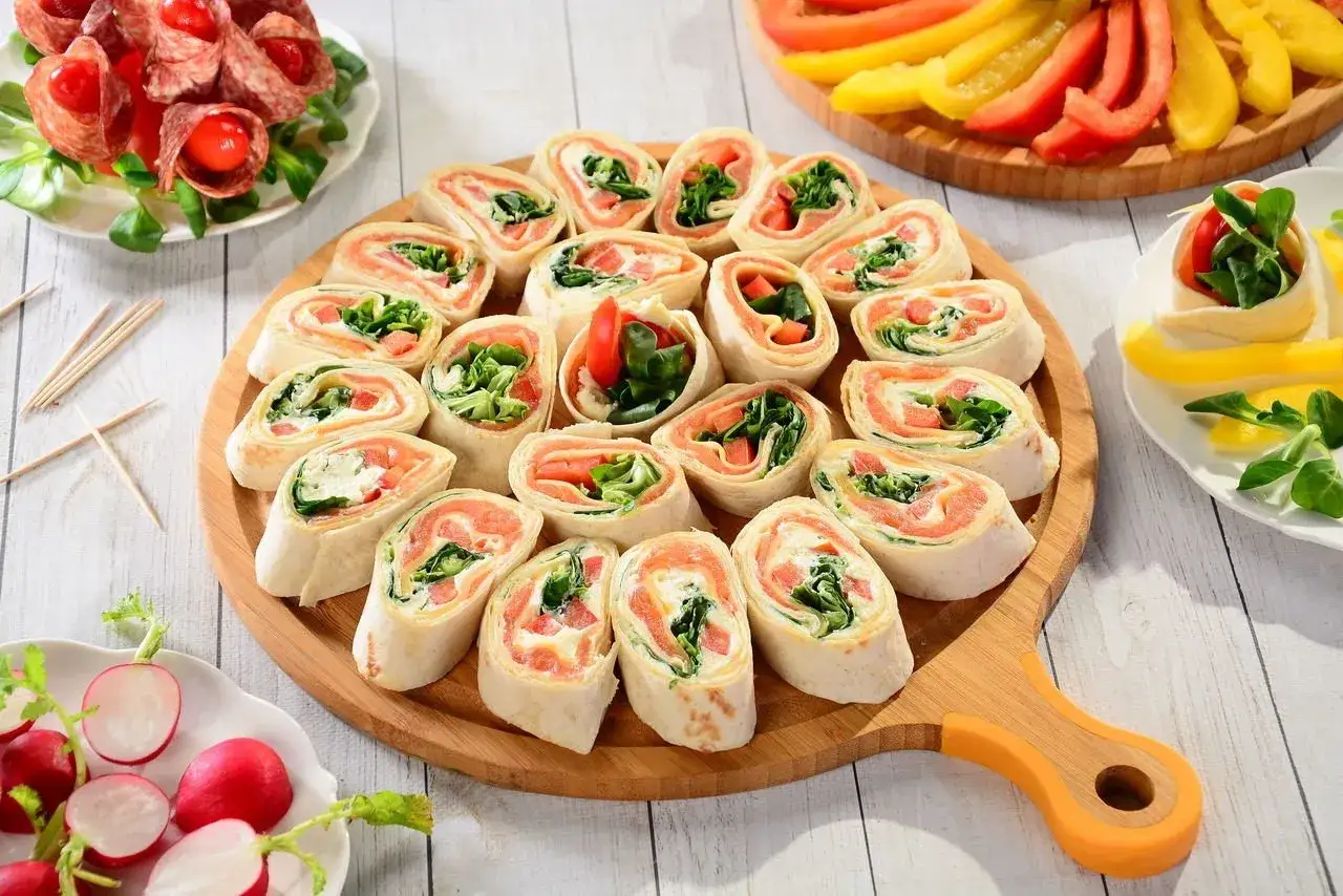Roladki z tortilli z łososiem, kremowym serkiem i rukolą, ułożone na drewnianej desce. Obok talerzyk z salami i pomidorkami, oraz kolorowe papryki.