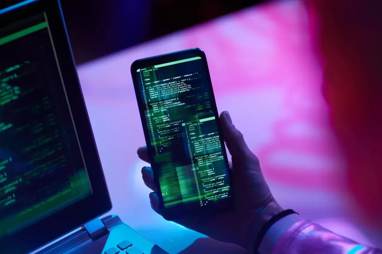 Ręka trzyma smartfon z kodem, obok laptop. Jak sprawdzić czy ktoś ma dostęp do mojego telefonu?