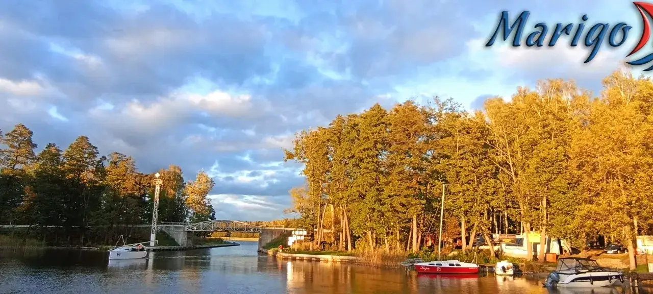 Mazury słyną z malowniczych kanałów, mostów i żaglówek. Słońce oświetla drzewa i wodę, tworząc spokojny krajobraz.
