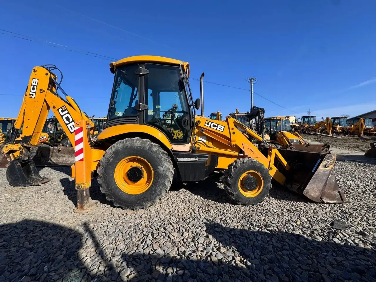 Żółta koparko-ładowarka JCB na placu z innymi maszynami. Zastanawiasz się, jaką koparko ładowarkę kupić? Ta może być dobrym wyborem.