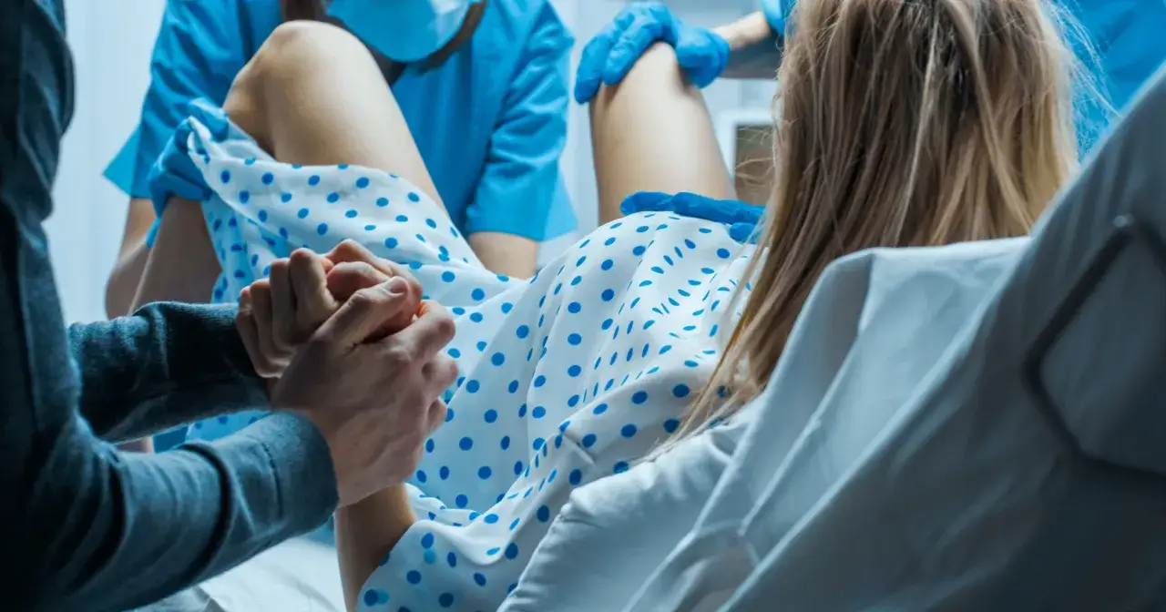 Wsparcie dla rodzącej kobiety, która trzyma za rękę partnera. W tle widać personel medyczny i biało-niebieski materiał, jak balonik do porodu.
