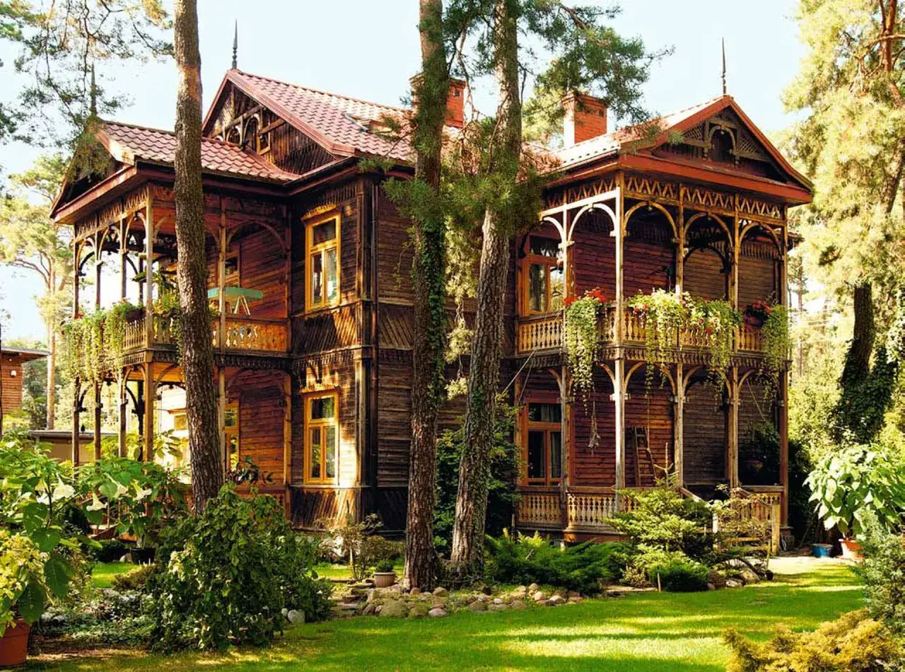 Drewniany dom w stylu świdermajer, z werandami i bogatymi zdobieniami, otoczony zielenią.