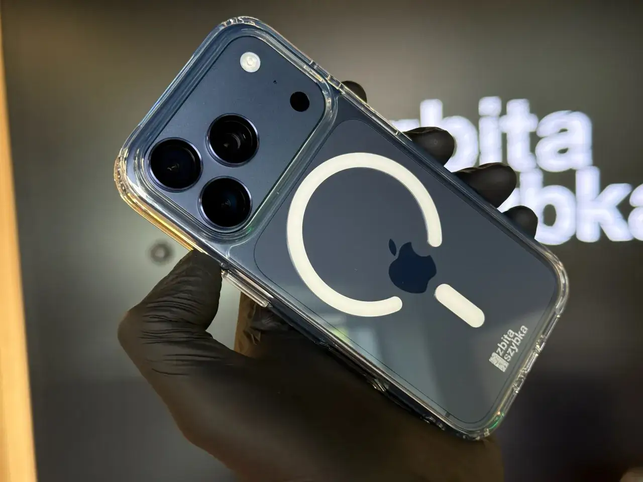 Ręka w czarnej rękawiczce trzyma iPhone'a w przezroczystym etui z widocznym modułem aparat&oacute;w i logo MagSafe. Dowiedz się, jak założyć szkło na aparat w telefonie.