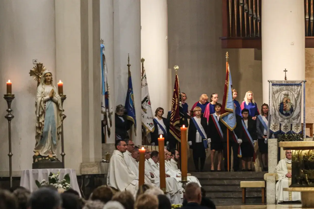 Uroczystość w kościele z figurą Matki Bożej, zapalonymi świecami i sztandarami. Wśród zgromadzonych wiernych, przykłady intencji mszalnych mogą być odczytywane.