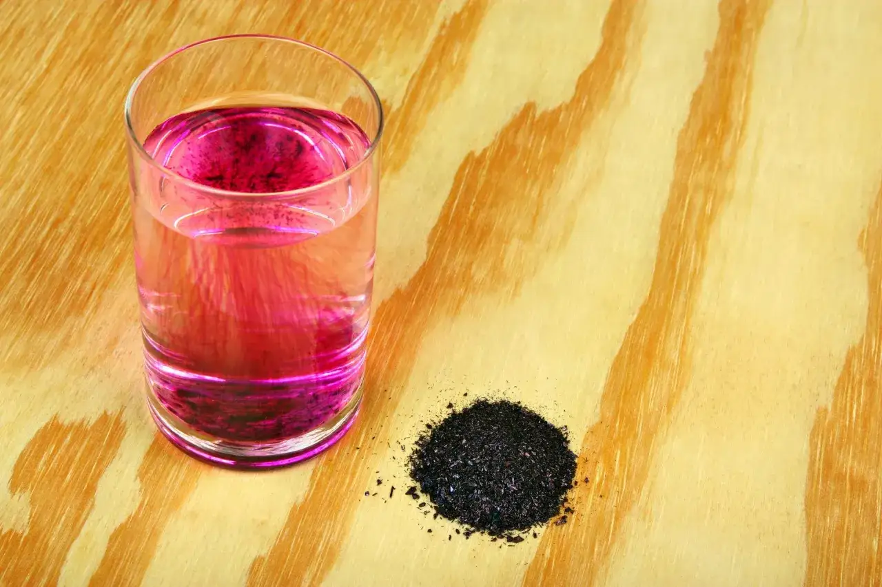 Szklanka z wodą zabarwioną na r&oacute;żowo przez nadmanganian potasu, obok kupka ciemnych kryształk&oacute;w.