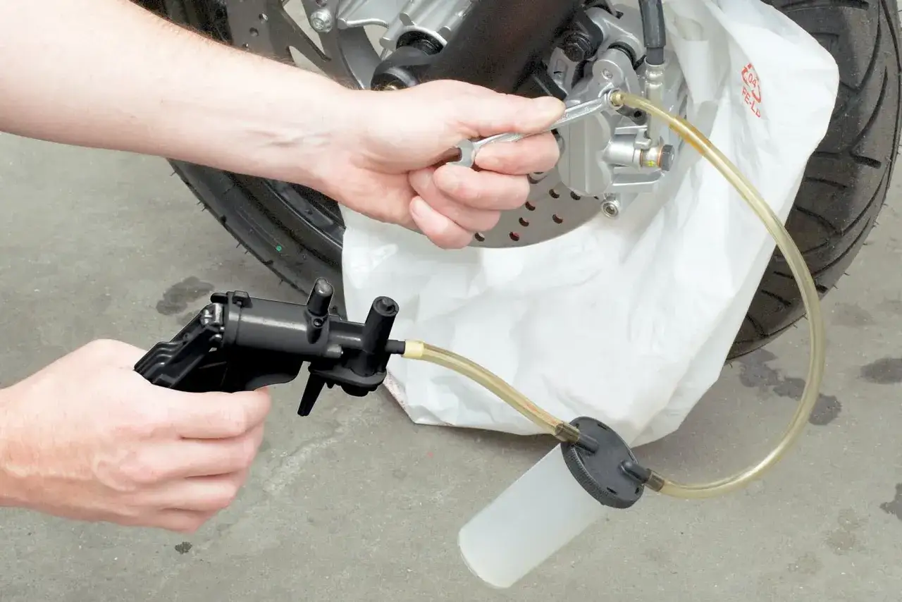 Ręce mechanika dokonują wymiany płynu hamulcowego w motocyklu, używając specjalnego narzędzia do odpowietrzania.