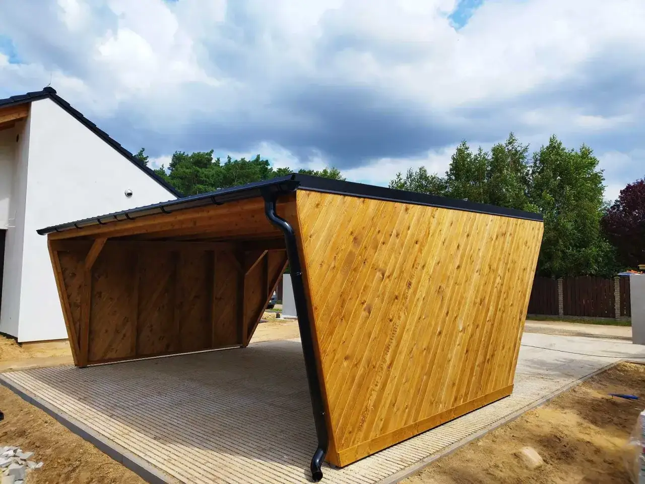 Drewniana wiata, która nie wymaga pozwolenia na budowę ani zgłoszenia, z dachem i rynnami, stoi na betonowym podłożu.