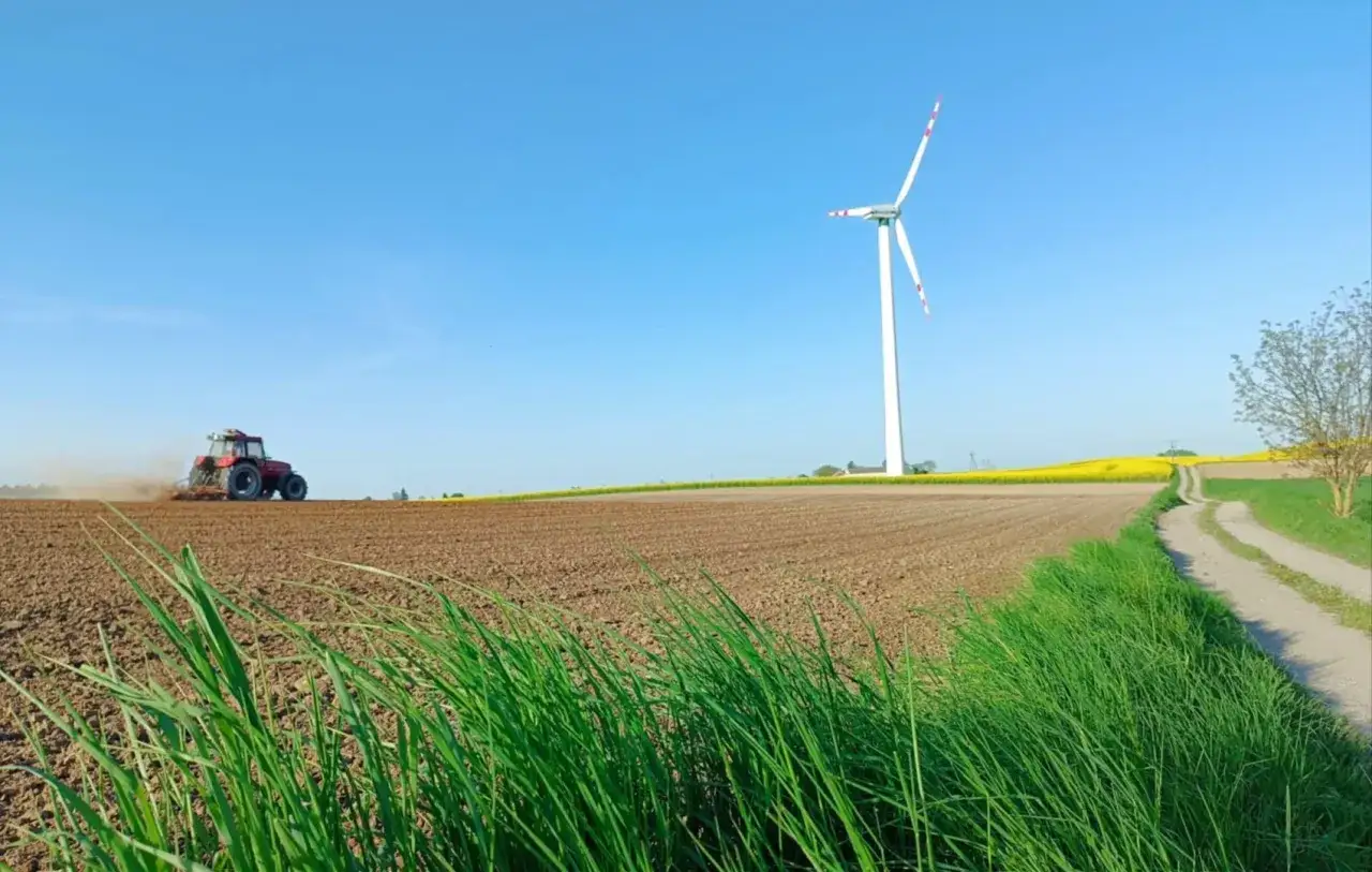 Czerwony traktor orze pole, a w tle wiatrak na polu pod błękitnym niebem.