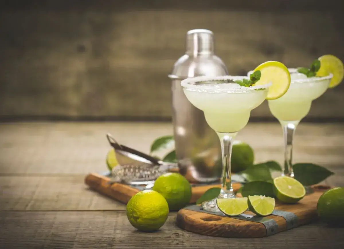 Dwa orzeźwiające drinki Margarita z solą na brzegach kieliszków, udekorowane plasterkami limonki i listkami mięty. Obok shaker i świeże limonki, idealne do przygotowania Margarita przepis.