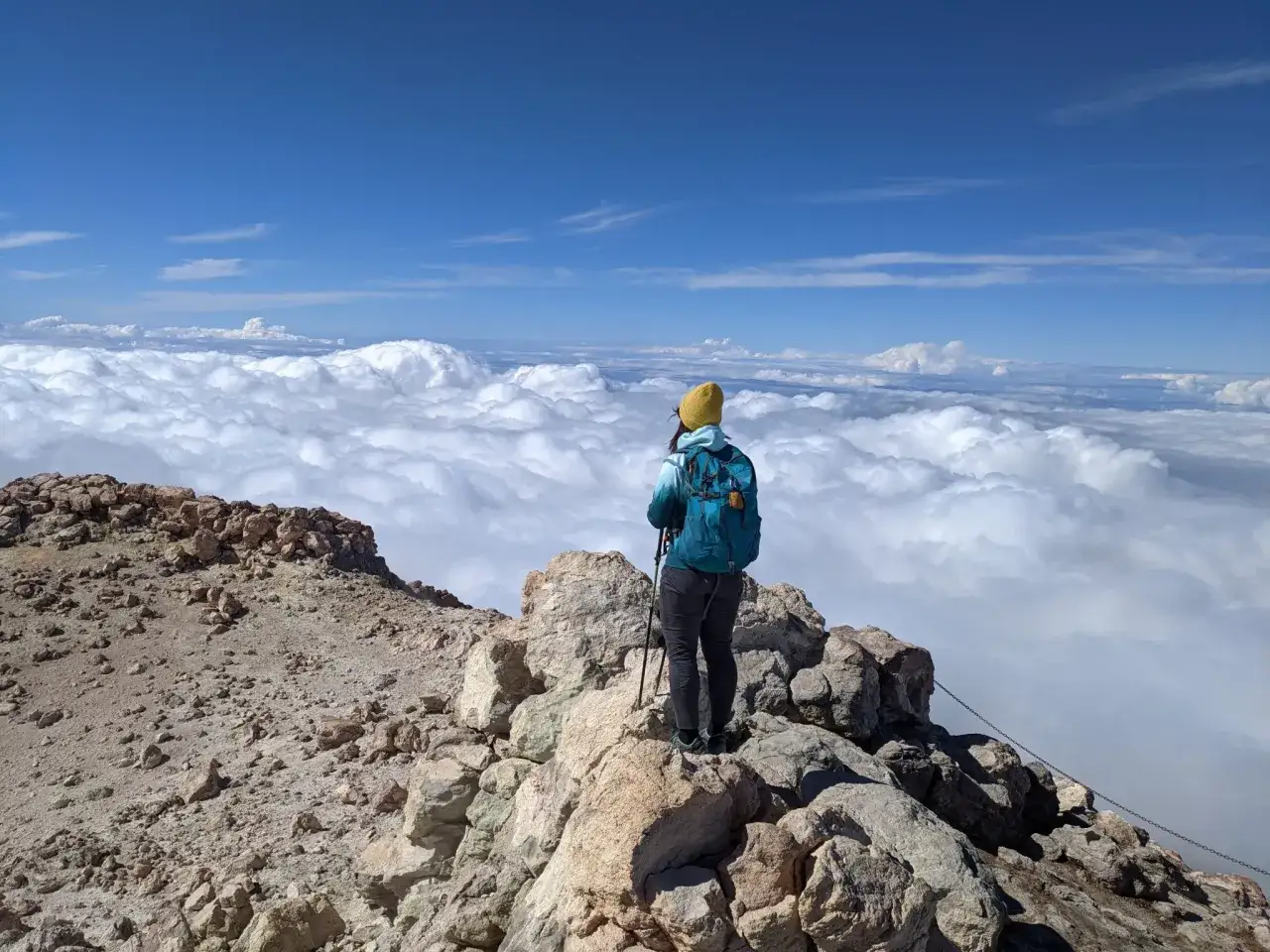 Widok z góry na chmury podczas wejścia na Teide. Turystka w żółtej czapce i niebieskim plecaku stoi na skalistym szczycie.