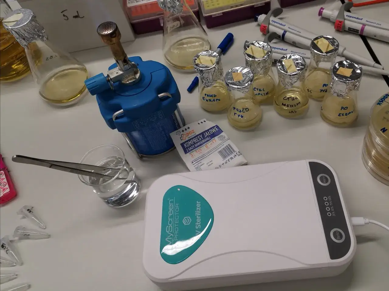 Czy odkażanie telefonu promieniami UV to skuteczna metoda na bakterie? Na stole laboratoryjnym widzimy sterylizator UV, probówki z pożywkami i palnik.