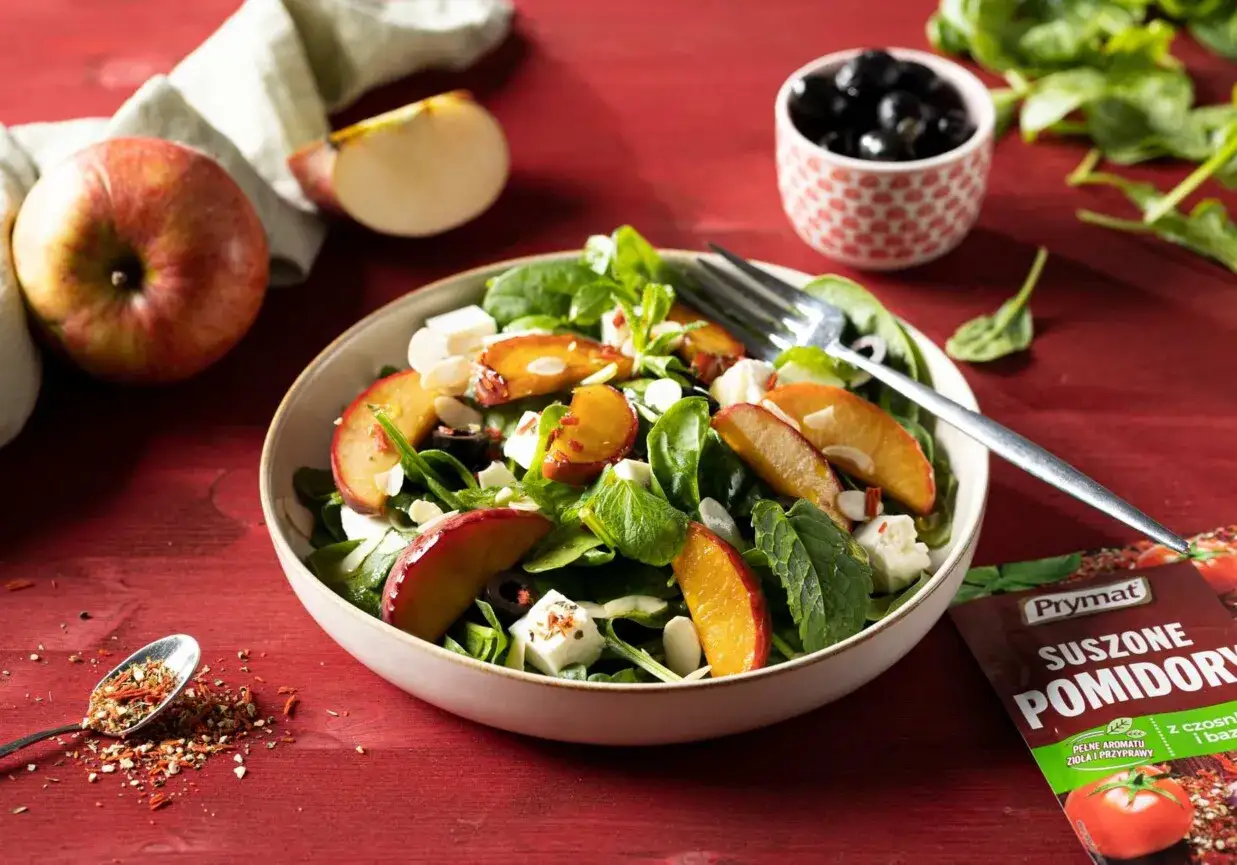 Pyszna sałatka ze świeżego szpinaku z kawałkami jabłka, serem feta i migdałami. Obok jabłko i oliwki.