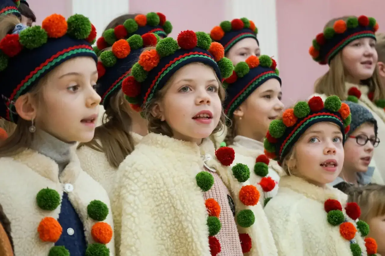 Dzieci w strojach ludowych, śpiewające kolędę. Ich czapki zdobią pompony, jakby nawiązując do czasów, kiedy powstała kolęda 