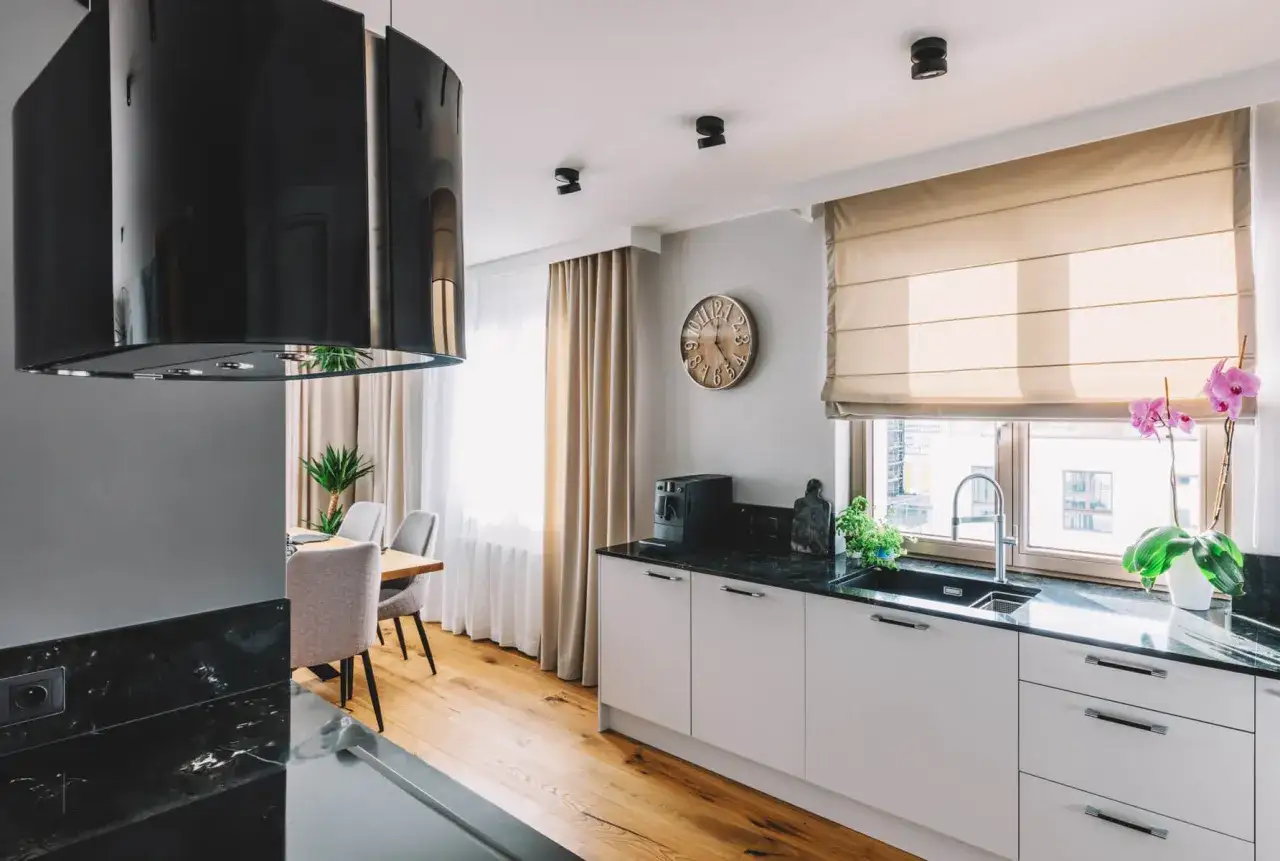 Nowoczesna kuchnia z czarnym okapem i halogenami w suficie podwieszanym. Obok okna zegar i ekspres do kawy.