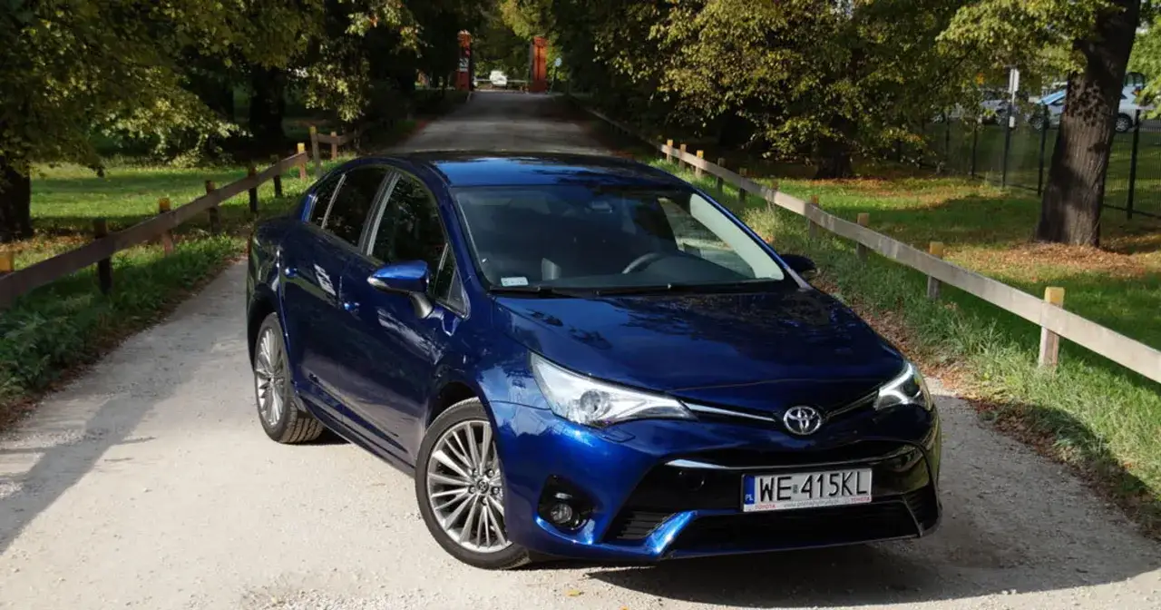 Błękitna Toyota Avensis na żwirowej drodze, otoczona zielenią i drzewami.