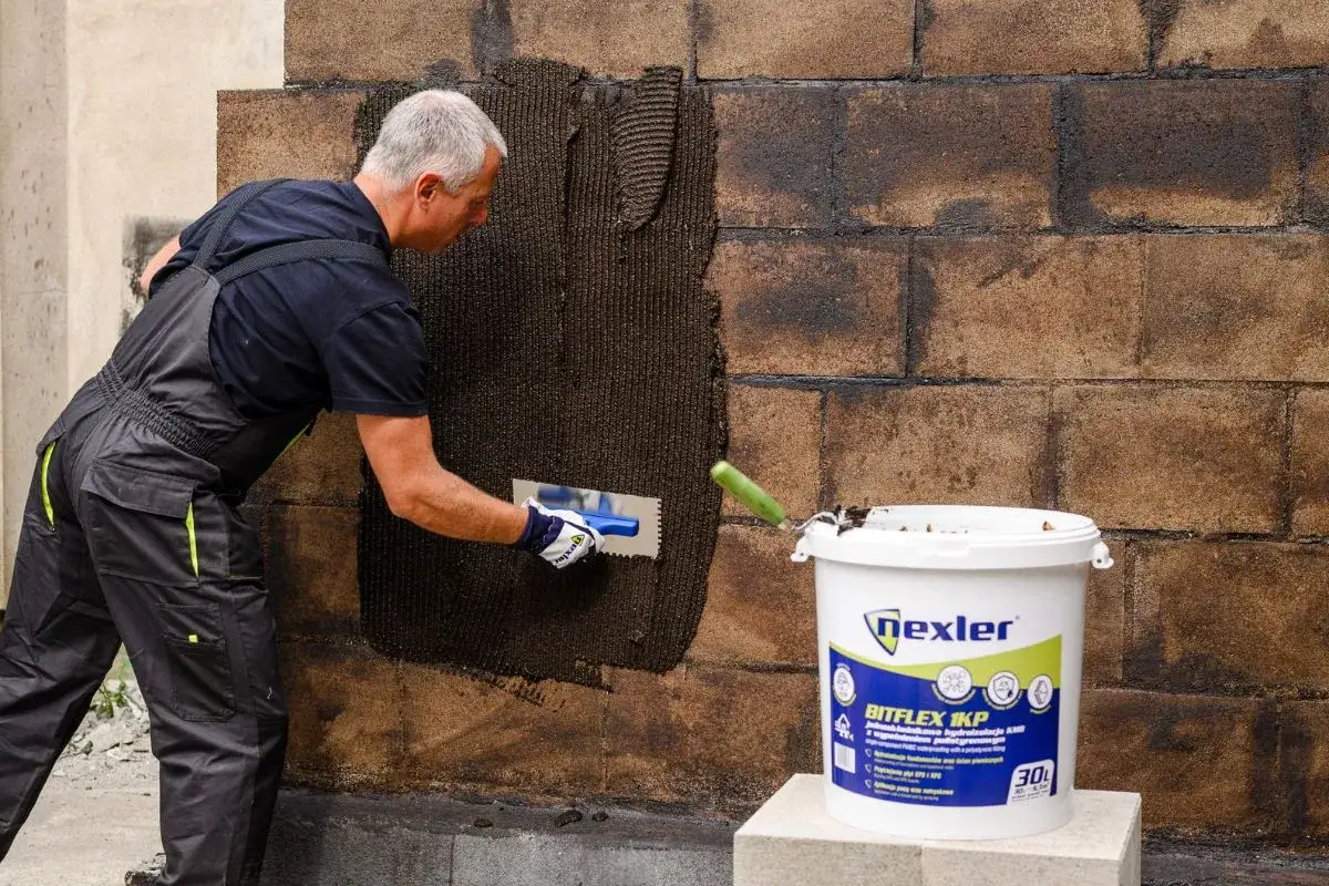 Budowlaniec nakłada masę na ścianę, wykonując hydroizolację ławy fundamentowej. Obok stoi wiadro z produktem.